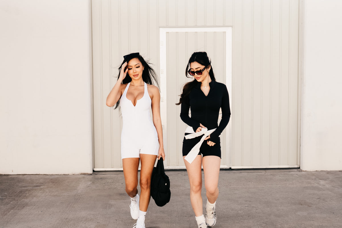 Two women walking beside a pale wall, wearing white and black activewear.